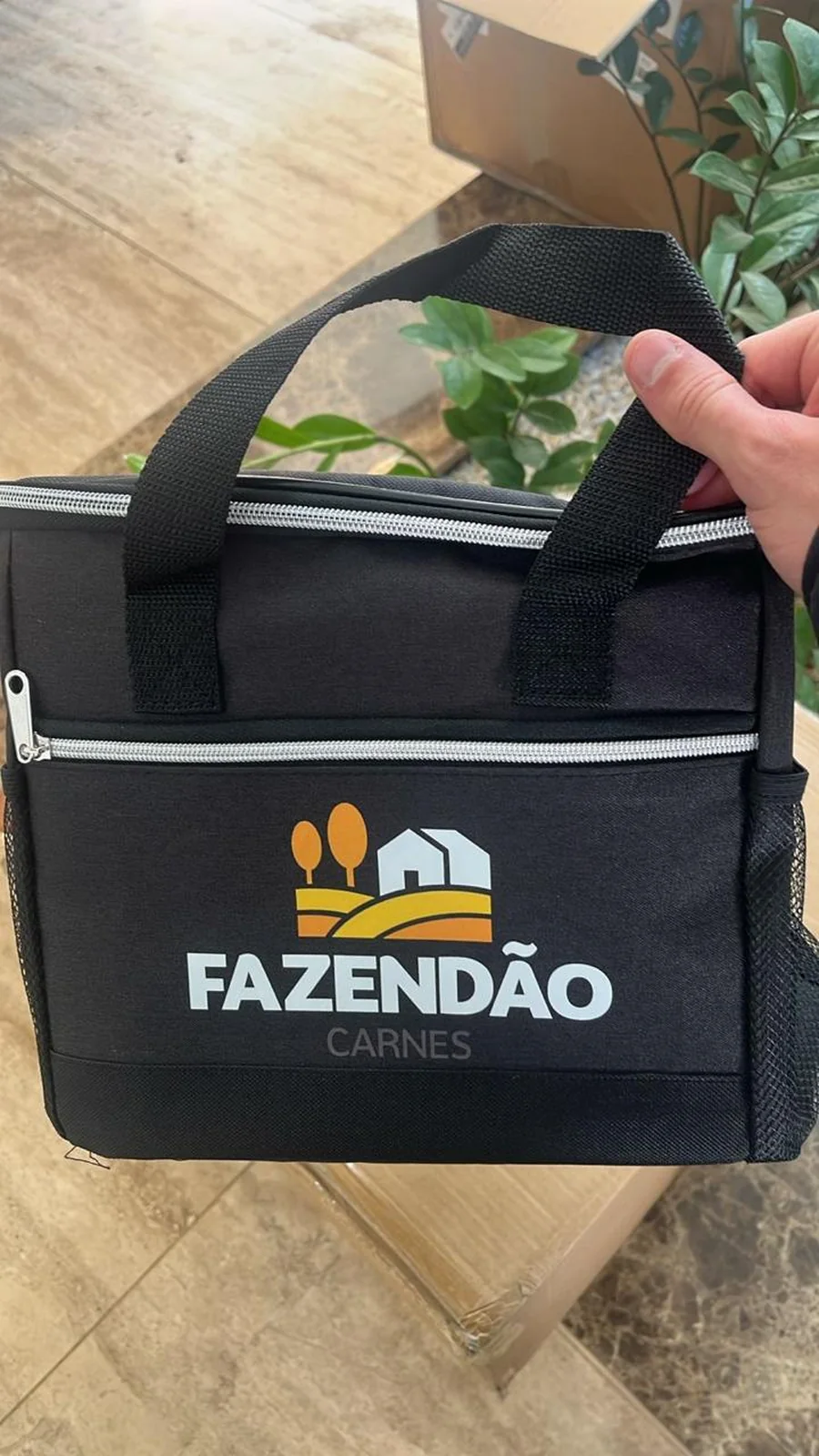 Bolsa térmica preta com alças, zíper prateado e a marca Fazendão Carnes, segurada sobre caixas