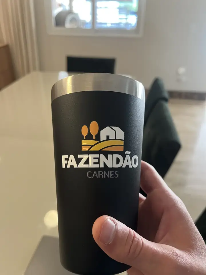 Mão segurando um copo térmico preto com a marca Fazendão Carnes, em uma sala de escritório