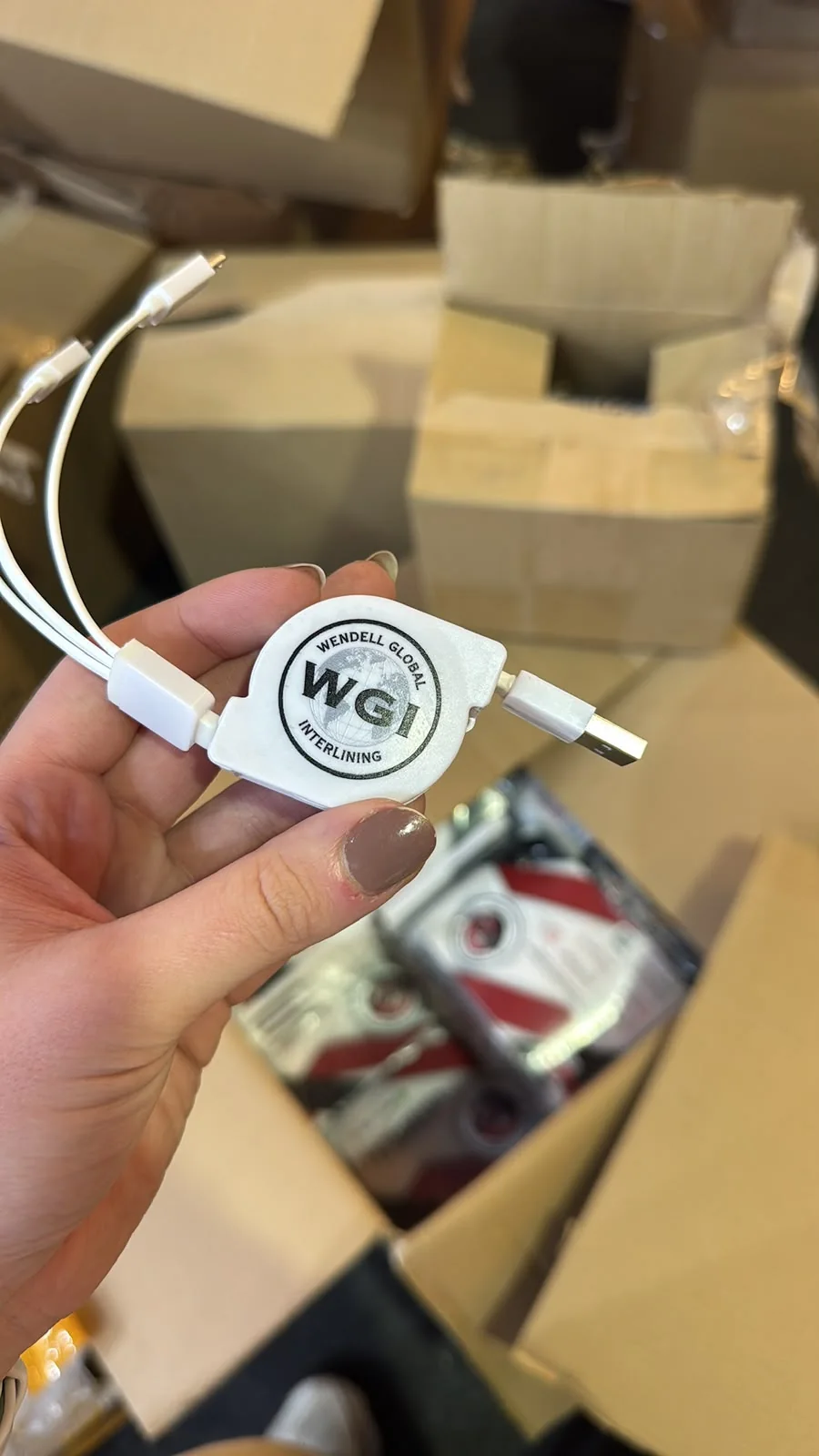 Mão segurando um cabo USB retrátil branco com o logotipo WGI, entre caixas de um estoque